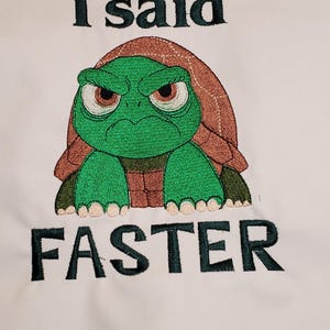 Puede incluir: Diseño bordado sobre tela blanca con una tortuga verde gruñona con caparazón marrón. El texto "I said FASTER" está bordado en verde oscuro, transmitiendo urgencia y humor.