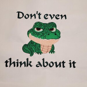 May include: Embroidered design featuring a green alligator with an angry expression. The text "Don't even think about it" is embroidered above and below the alligator. The design is on a white background.