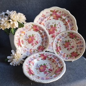 May include: A set of four ceramic dishes featuring a cream-colored base with a repeating floral pattern in shades of pink, red, and green. Each piece has a scalloped, fluted edge. The set includes one large platter, two plates, and one bowl.