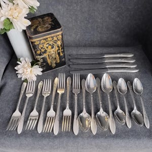 Puede incluir: Una colección de cubiertos antiguos, que incluye tenedores, cucharas y cuchillos, dispuestos sobre una superficie gris. También hay una lata negra decorativa con detalles dorados y un jarrón blanco con flores. Los cubiertos parecen ser de plata.