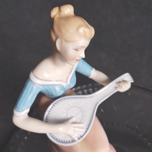 May include: A porcelain figurine of a woman playing a white mandolin. The woman wears a light blue dress with a pink skirt. Her hair is styled in a bun. The mandolin has a detailed design.