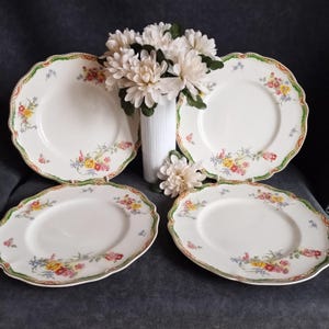Może przedstawiać: Zestaw czterech białych talerzy ceramicznych z falistymi brzegami, ozdobionych zielono-pomarańczową obwódką oraz delikatnym kwiatowym wzorem w odcieniach różu, żółci i błękitu, ustawionych wokół centralnego białego, żebrowanego wazonu.