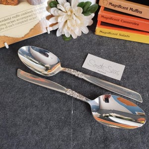 Può includere: Due cucchiai da portata argentati con manici decorati. I cucchiai sono riflettenti, con un motivo floreale sui manici. È visibile un piccolo biglietto con "South Seas Design". Lo sfondo include libri di cucina e un fiore bianco.