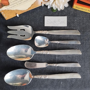 Può includere: Un set di posate d'argento da portata, tra cui una forchetta, cucchiai e un coltello da burro, disposte su una superficie grigio scuro. Le posate hanno manici ornati e sono di South Seas Design. Un libro di ricette e una scatola di sale sono sullo sfondo.
