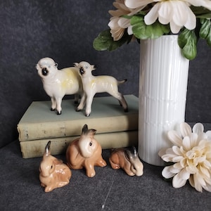 Peut inclure: Ensemble de figurines en céramique représentant deux moutons et trois lapins. Les moutons sont blancs avec des détails noirs, tandis que les lapins sont bruns et beiges. Les figurines sont disposées avec un vase blanc et des fleurs en arrière-plan.