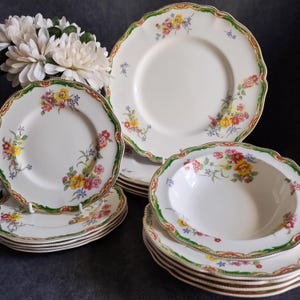 May include: A set of cream-coloured ceramic dinnerware featuring a scalloped edge with a green border and a floral pattern of red, yellow, and blue flowers. The collection includes stacked dinner plates, salad plates, and shallow soup bowls.