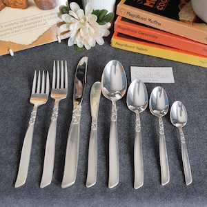 May include: A silver cutlery set with decorative handles, comprising forks, knives, and spoons. The set is displayed on a grey surface, with cookbooks and a flower in the background. The text "South Seas Design" is visible.