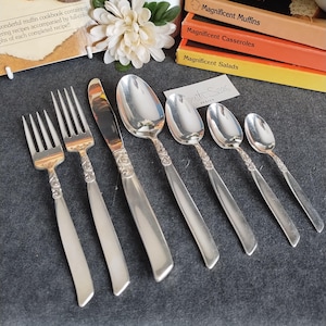 Puede incluir: Un juego de cubiertos plateados, que incluye tenedores, cuchillos y cucharas, dispuestos sobre una superficie gris oscuro. Los mangos presentan un diseño ornamentado. Libros de cocina con títulos como "Magnificent Muffins" están en el fondo.