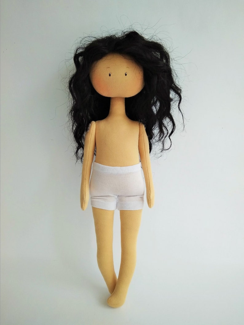 May include: A handmade doll with long, curly black hair, a beige body, and white underwear. The doll has a simple face with black eyes and a small, pink nose.