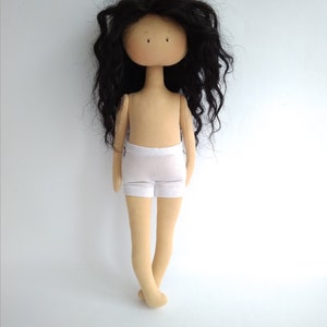 May include: A handmade doll with long, black, curly hair. The doll is wearing a white, cotton undergarment. The doll has a beige body and a neutral facial expression.