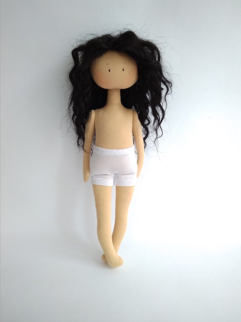 May include: A handmade doll with long, black, curly hair. The doll is wearing a white, cotton undergarment. The doll has a beige body and a neutral facial expression.