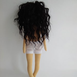 May include: A handmade doll with long, curly black hair, wearing a white skirt and standing on a white background.