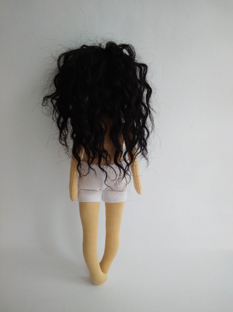 May include: A handmade doll with long, curly black hair, wearing a white skirt and standing on a white background.