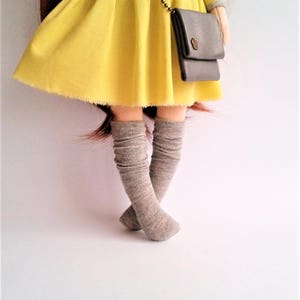 May include: A doll wearing a yellow dress with a gray striped top and a gray purse. The doll is wearing gray knee-high socks.