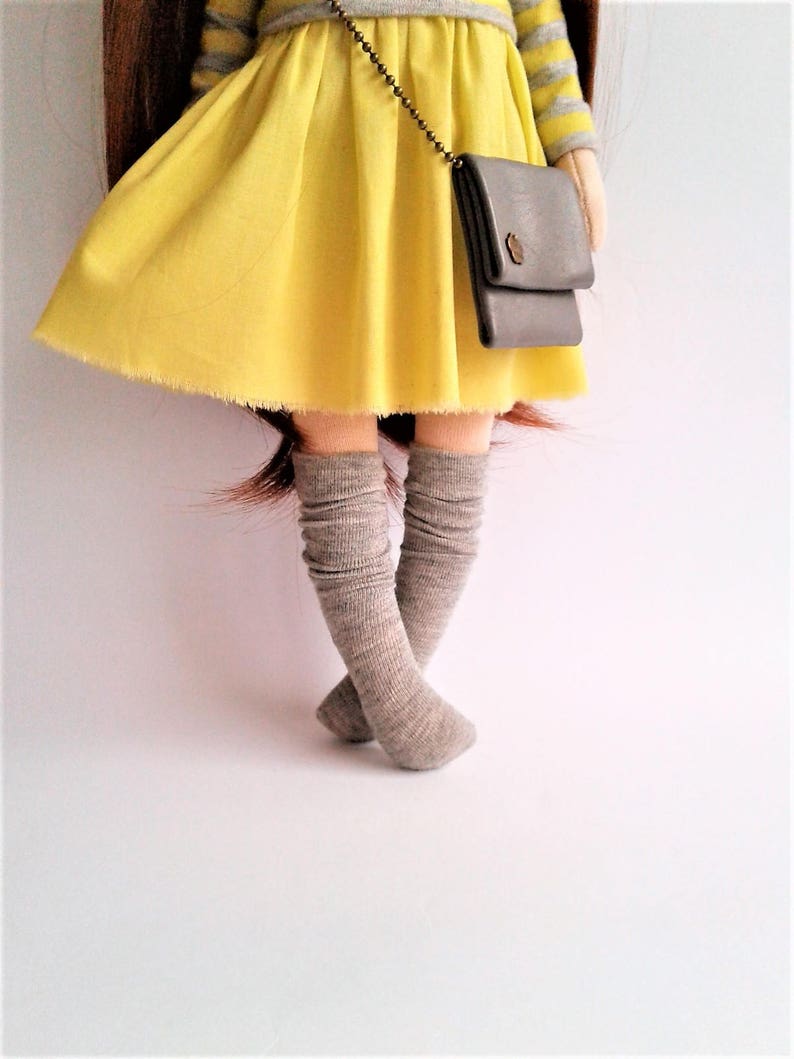 May include: A doll wearing a yellow dress with a gray striped top and a gray purse. The doll is wearing gray knee-high socks.