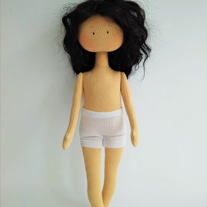 May include: A handmade doll with long black curly hair, a beige body, and white underwear. The doll is standing on a white background.