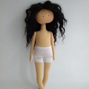 May include: A handmade doll with long, curly black hair, a beige body, and white underwear. The doll has a simple face with black eyes and a beige complexion.