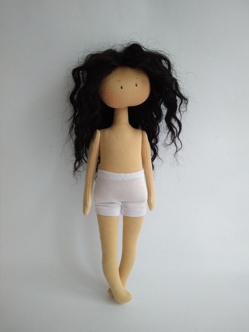 May include: A handmade doll with long, curly black hair, a beige body, and white underwear. The doll has a simple face with black eyes and a beige complexion.