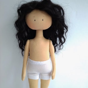 May include: A handmade doll with long, curly black hair, a beige body, and white shorts. The doll has a simple, round face with painted eyes.