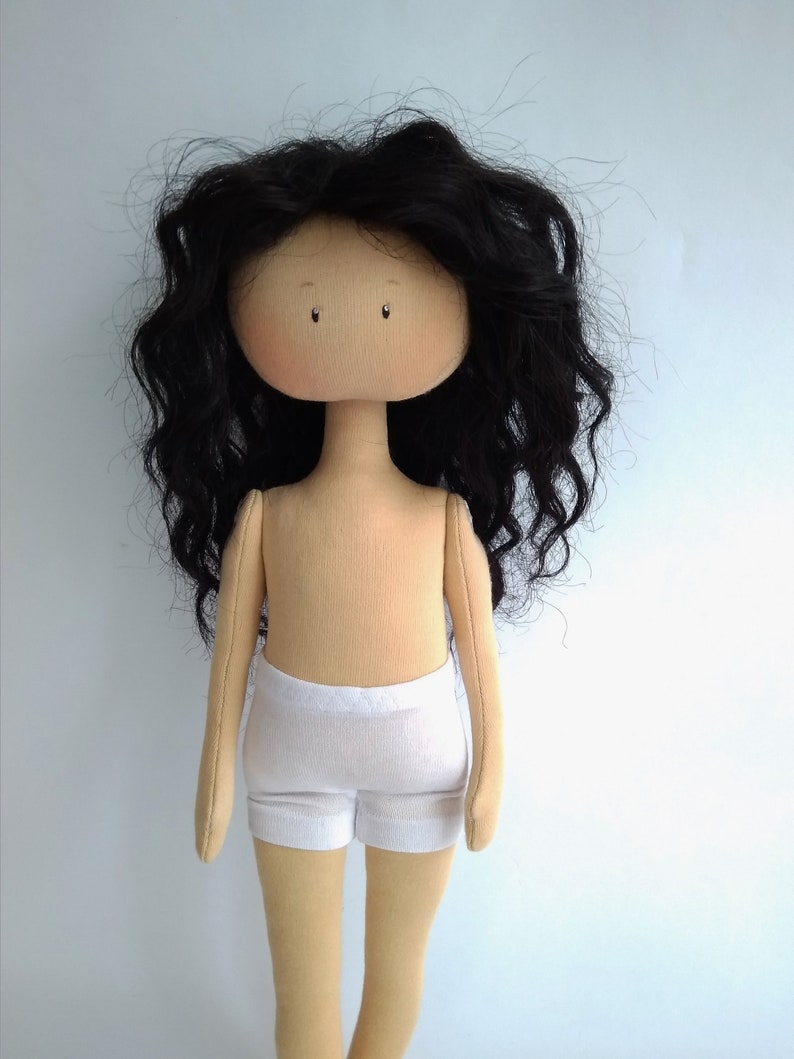 May include: A handmade doll with long, curly black hair, a beige body, and white shorts. The doll has a simple, round face with painted eyes.
