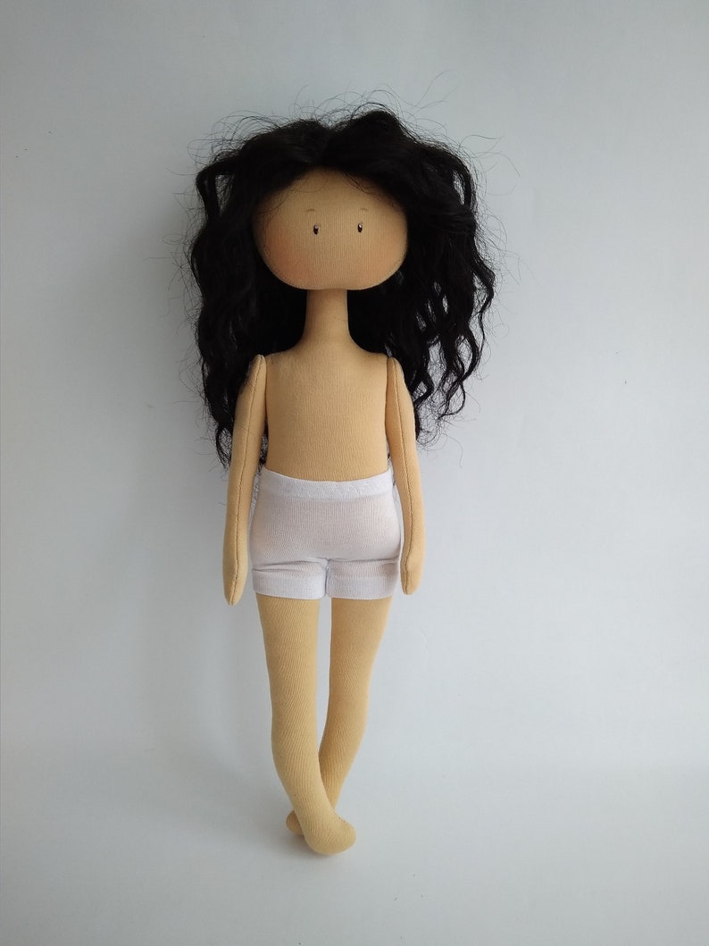 May include: A handmade doll with long black curly hair, a beige body, and white underwear. The doll has a simple face with brown eyes.