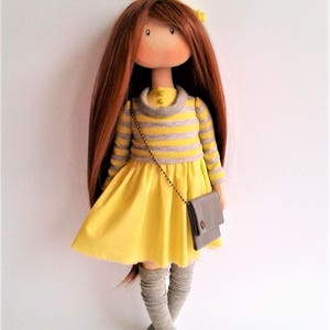 May include: A handmade doll with long brown hair, wearing a yellow dress with grey stripes, grey knee-high socks, and a small grey purse.