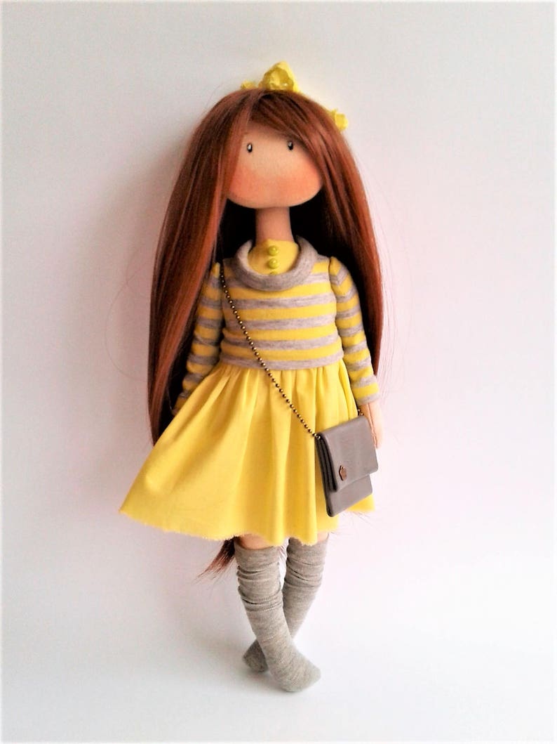 May include: A handmade doll with long brown hair, wearing a yellow dress with grey stripes, grey knee-high socks, and a small grey purse.
