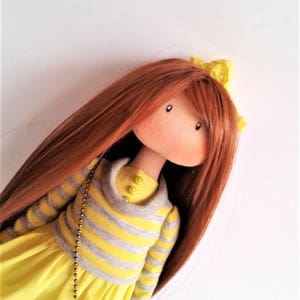 May include: A handmade doll with long, auburn hair, wearing a yellow dress with a gray and yellow striped sweater. The doll has a yellow headband and green buttons on the sweater.