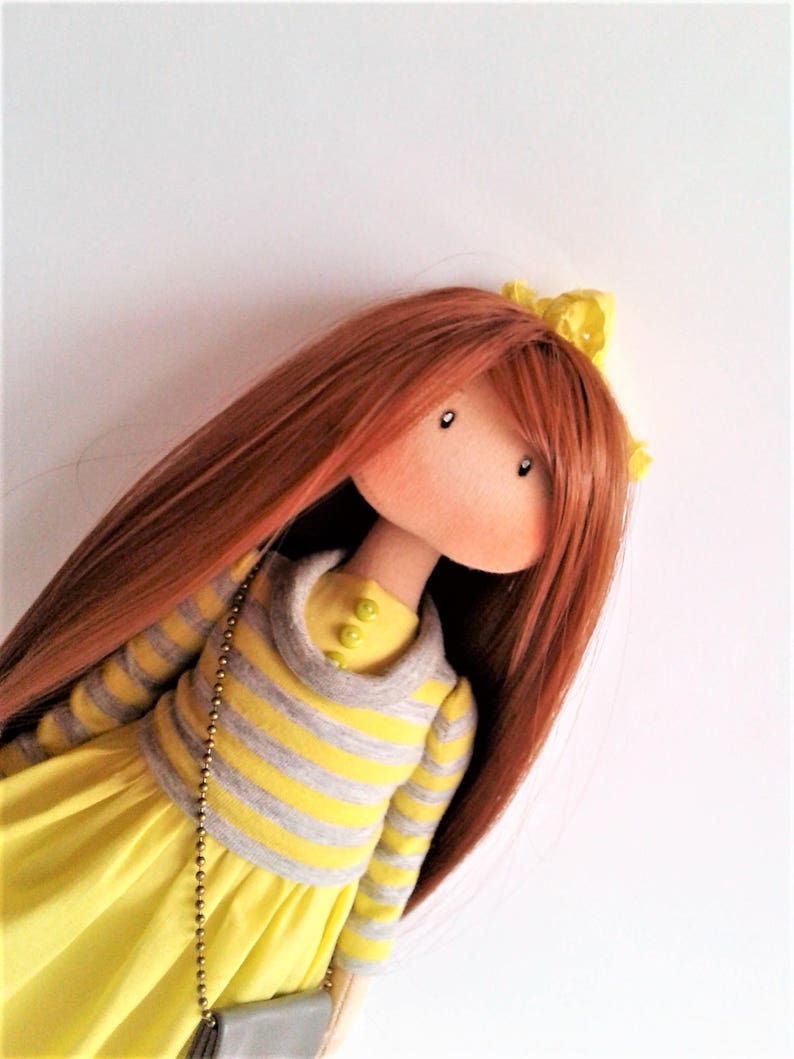 May include: A handmade doll with long, auburn hair, wearing a yellow dress with a gray and yellow striped sweater. The doll has a yellow headband and green buttons on the sweater.