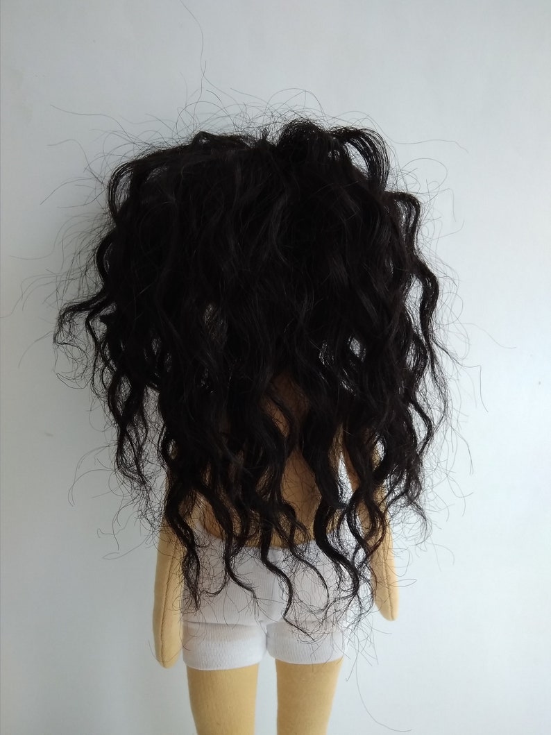 May include: A handmade doll with long, curly black hair and a white fabric skirt. The doll is standing on a white background.