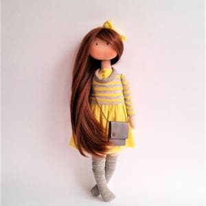 May include: A handmade doll with long brown hair, wearing a yellow and gray striped shirt, a yellow skirt, gray knee-high socks, and a small gray purse.