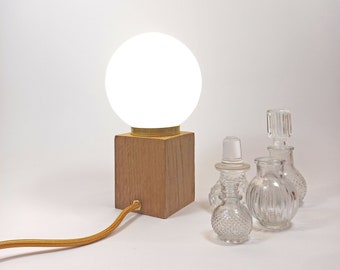 Puede incluir: Una lámpara de globo blanco con una base de madera y un cable dorado. La lámpara está sobre una superficie blanca con tres botellas de vidrio transparente.