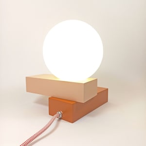 May include: A modern table lamp with a white globe shade and a two-tiered base. The base is made of wood and is painted in shades of orange and beige.