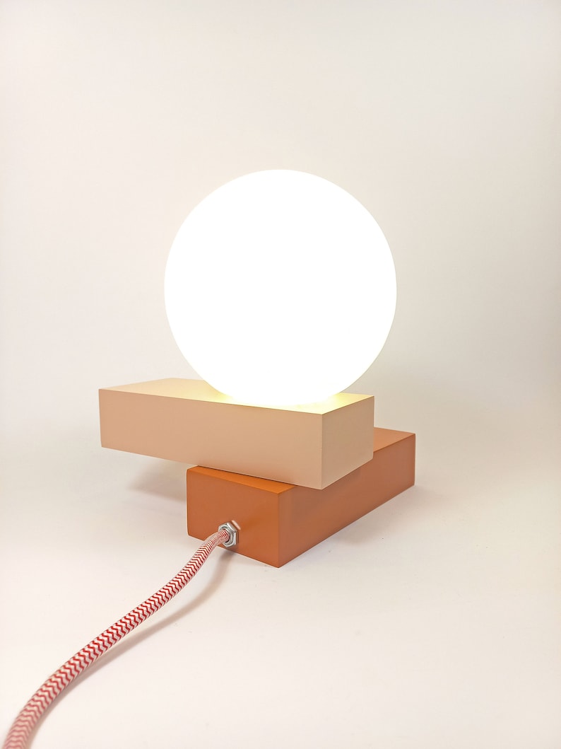 May include: A modern table lamp with a white globe shade and a two-tiered base. The base is made of wood and is painted in shades of orange and beige.