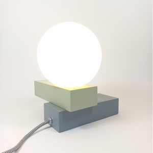 May include: A modern table lamp with a white globe shade and a geometric base made of two rectangular blocks. The top block is light green and the bottom block is light blue.