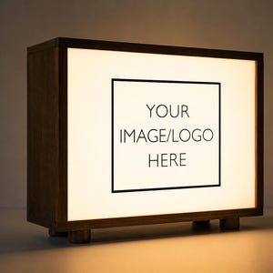 Può includere: Una scatola luminosa rettangolare con una cornice in legno marrone scuro e quattro piccoli piedini cilindrici. Il pannello frontale è illuminato e presenta un bordo rettangolare nero con il testo YOUR IMAGE/LOGO HERE centrato in nero.