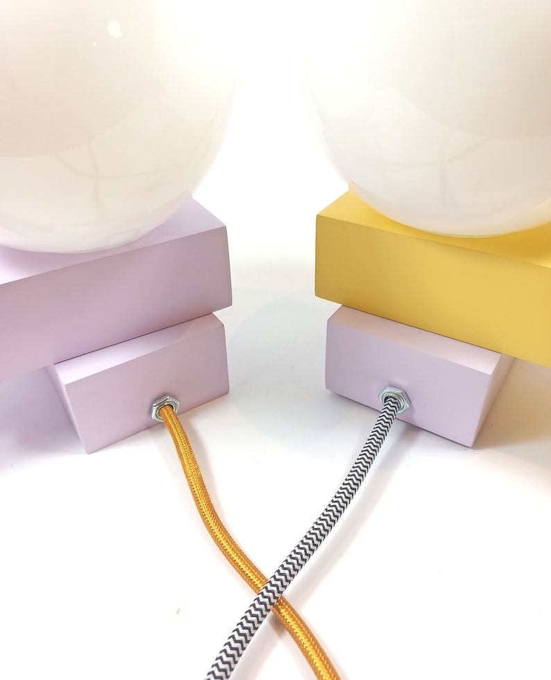 May include: Two table lamps with white globe shades, one with a lavender base and the other with a yellow base. The lamps have black and white patterned cords and gold cords.