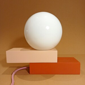 May include: A white globe light fixture sits on top of two rectangular blocks. The top block is light pink and the bottom block is orange. A white cord with red stripes is attached to the base.
