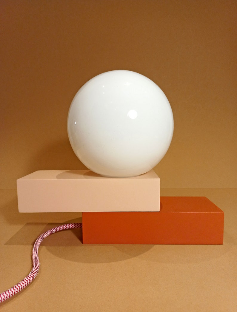 May include: A white globe light fixture sits on top of two rectangular blocks. The top block is light pink and the bottom block is orange. A white cord with red stripes is attached to the base.