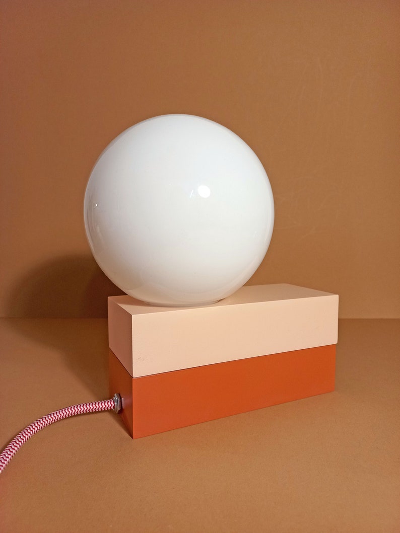 May include: A white globe lamp sits on top of a two-toned rectangular base. The base is peach on top and a darker orange on the bottom.