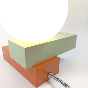 May include: A modern table lamp with a white orb shade and a two-tiered base. The base is made of wood and painted in a light green and orange color. The lamp has a black and white cord.