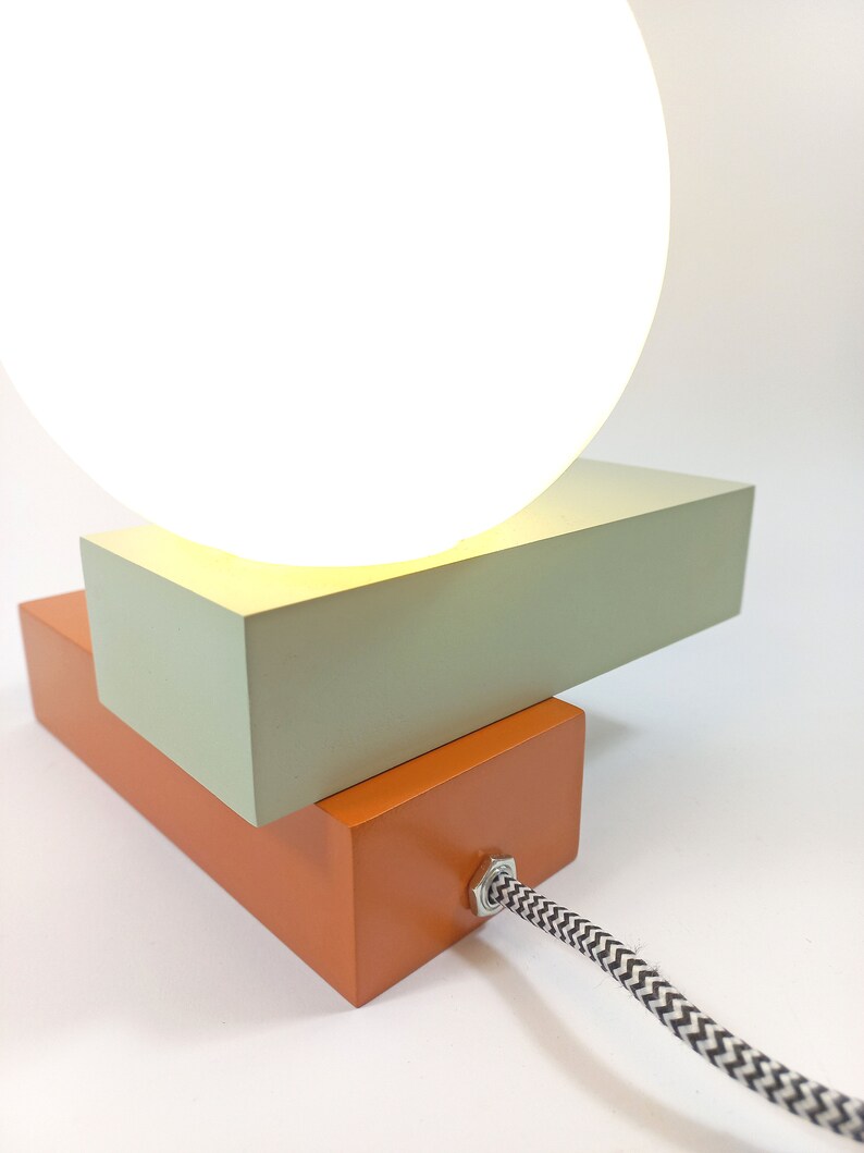 May include: A modern table lamp with a white orb shade and a two-tiered base. The base is made of wood and painted in a light green and orange color. The lamp has a black and white cord.