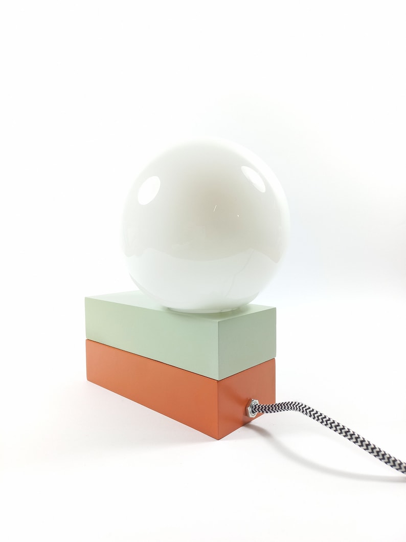 May include: A table lamp with a white globe shade and a two-toned base. The base is made of wood and is painted in shades of green and orange.