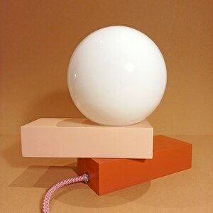 May include: A modern table lamp with a white globe shade and a two-toned base. The base is made of two rectangular blocks, one peach and one orange.