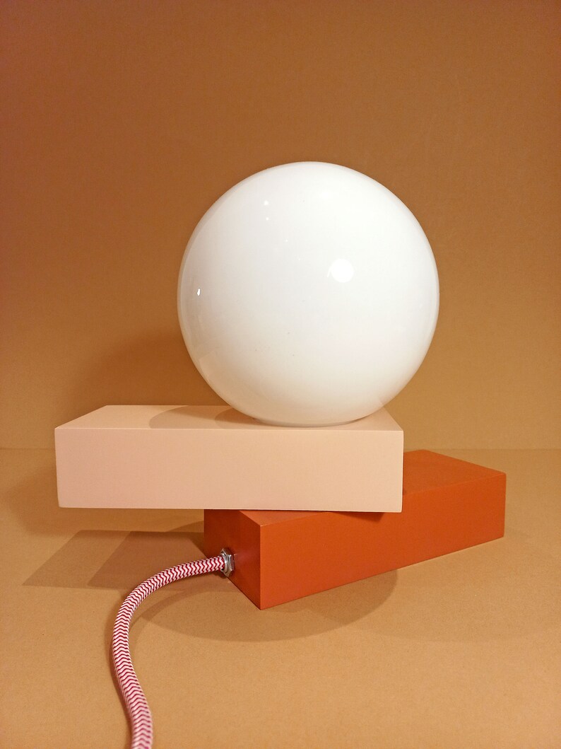 May include: A modern table lamp with a white globe shade and a two-toned base. The base is made of two rectangular blocks, one peach and one orange.
