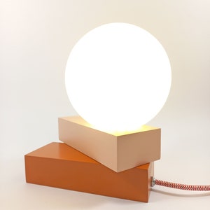 May include: A modern table lamp with a white globe shade and a two-toned base. The base is made of wood and is painted in shades of orange and pink.