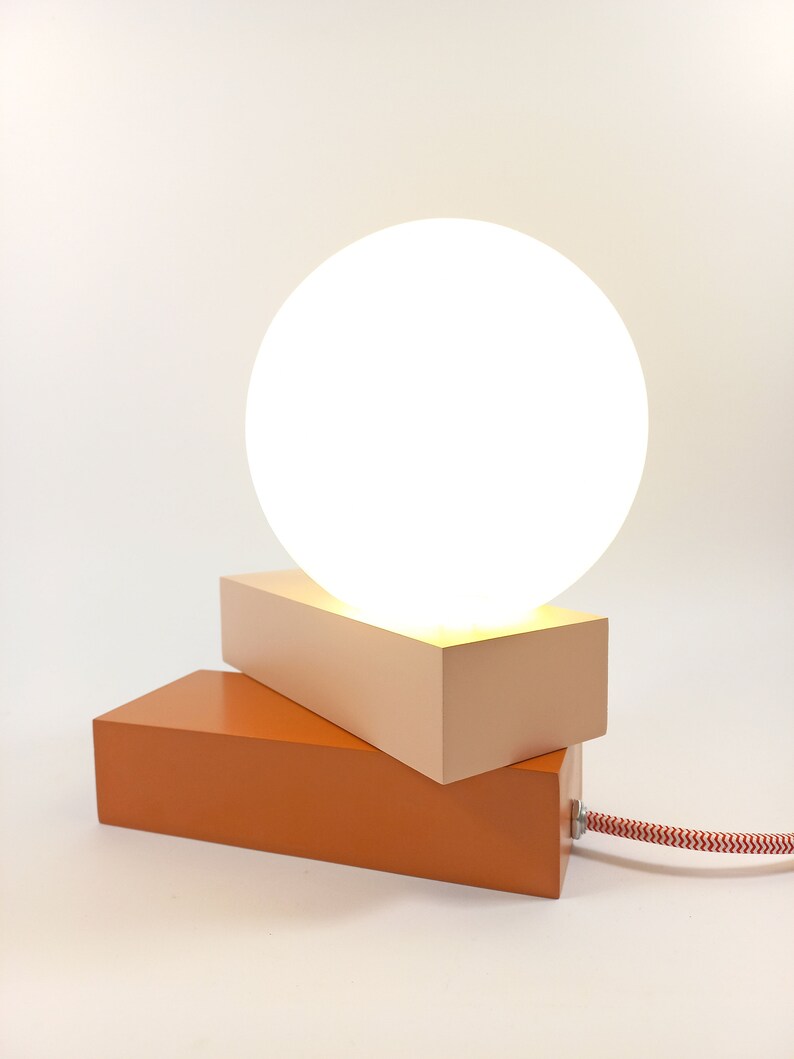 May include: A modern table lamp with a white globe shade and a two-toned base. The base is made of wood and is painted in shades of orange and pink.