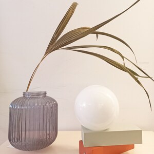 May include: A gray ribbed glass vase with a dried palm frond, a white globe lamp on top of two stacked rectangular blocks, one orange and one light green.