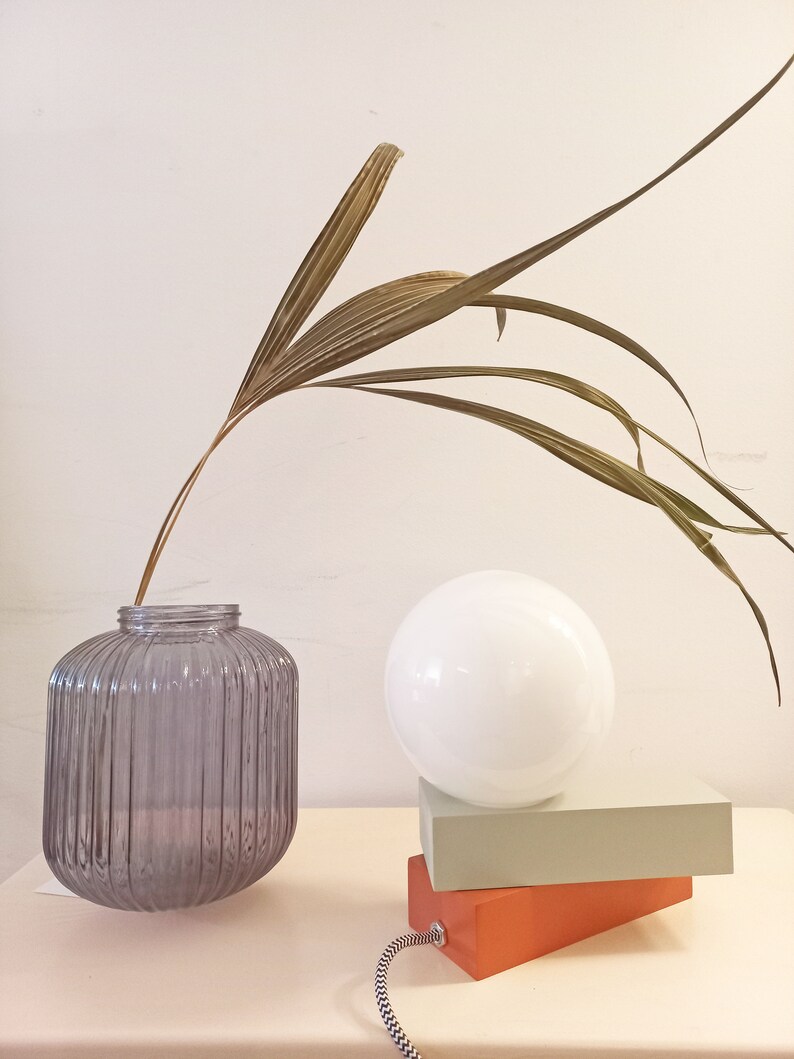 May include: A gray ribbed glass vase with a dried palm frond, a white globe lamp on top of two stacked rectangular blocks, one orange and one light green.