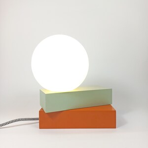 May include: A modern table lamp with a white globe shade and a stacked base of light green and orange rectangular blocks.